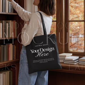 Pode incluir: Bolsa tote preta com o texto "Your Design Here" em uma fonte branca e elegante. A bolsa é carregada por uma pessoa em uma sala com uma estante e uma janela. O texto "LADYBUG MOCKS" também é visível.