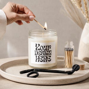 May include: A lit candle in a clear glass jar with a white label that reads "Your Design Here." A small glass bottle of matches, a black wick trimmer, and a beige tray are also present. The candle is on a beige surface.