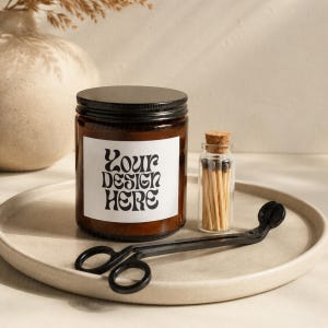 May include: A brown glass candle jar with a black lid and a white label that reads "Your Design Here." A small glass bottle with a cork stopper and matches, and a black wick trimmer sit on a round, beige tray.