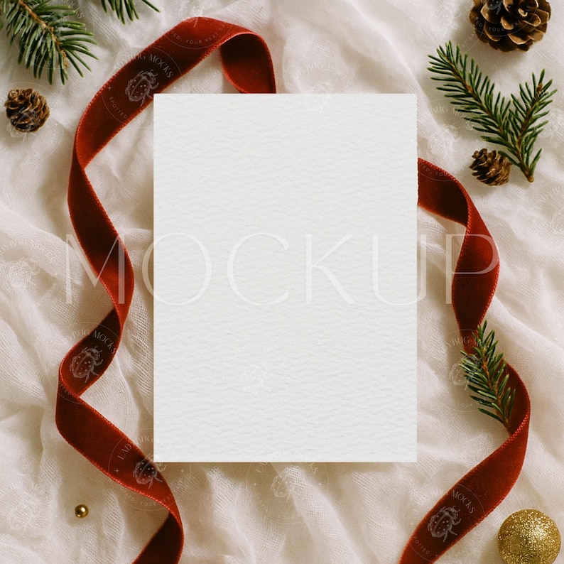 May include: A blank white card with a textured surface, surrounded by a red velvet ribbon, pine needles, pine cones, and a gold ornament. The ribbon has text that reads "LAB MOXAS". The scene is set on a white, textured fabric.