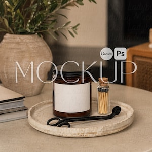 May include: A candle in a brown glass jar with a black lid and a blank white label sits on a round, light-colored tray. A small glass bottle of matches and a black wick trimmer are also on the tray. The word "MOCKUP" is displayed in the image.