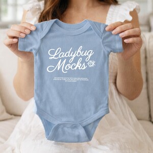Op de afbeelding: Een lichtblauwe babybody met de tekst "Ladybug Mocks" in een cursief lettertype. De body wordt door een persoon omhoog gehouden. De body heeft korte mouwen en een ronde halslijn. De tekst is wit.