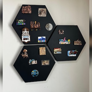 Op de afbeelding: Drie zwarte, zeshoekige magnetische borden met diverse souvenirmagneten. De magneten tonen bezienswaardigheden, steden en culturele symbolen van verschillende locaties. De borden zijn aan een witte muur bevestigd.