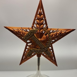 May include: A bronze-colored star-shaped sculpture with a hammer and sickle emblem. The star is mounted on a clear stand atop a silver base. The star's design includes wheat stalks and intricate detailing.