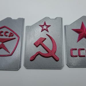 May include: Three gray rectangular objects with pink accents. Each features a different Soviet symbol: a star, a hammer and sickle, and a stylized emblem with the letters "CCCP".