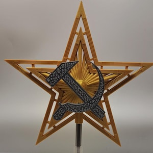 May include: A gold-colored star-shaped ornament featuring a hammer and sickle emblem. The star has a detailed, layered design with a sunburst pattern behind the emblem. The hammer and sickle are dark gray with a textured surface.