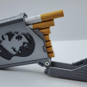 Könnte beinhalten: Graues Zigarettenetui mit schwarzem Grafikdesign. Das Etui ist geöffnet und zeigt mehrere Zigaretten. Das Etui besteht aus einem harten Material und hat einen Klappdeckel.