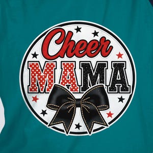 May include: Teal raglan-sleeve t-shirt with a circular graphic. The graphic reads "Cheer MAMA" in red and black lettering, with a black bow at the bottom. The shirt has navy blue sleeves and a white background.