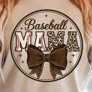 Puede incluir: Camiseta beige con un diseño circular con la frase "Baseball MAMA". El diseño incluye una pelota de béisbol, estampado de leopardo y un lazo marrón. La camiseta es de estilo casual.