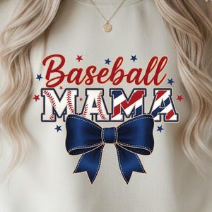 Puede incluir: Sudadera beige con la frase "Baseball MAMA" en rojo y azul, con un lazo azul. El texto está decorado con estrellas. La sudadera es de cuello redondo.