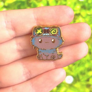May include: A gold-coloured enamel pin depicting a cartoon cat character wearing goggles. The cat is mainly blue and brown with a smiling face and small paws. The goggles have green and yellow accents.