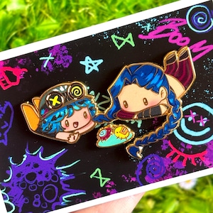 May include: Two enamel pins featuring cartoon characters with blue hair and gold accents. One pin depicts a character wearing a hat, the other a character with a long braid. The pins are on a black background with colourful graffiti.