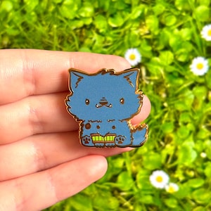 Puede incluir: Pin esmaltado de gato azul con ribete dorado. El gato está sentado con un libro delante. El pin se sostiene en una mano sobre un fondo de hierba verde y flores blancas.