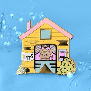 Könnte beinhalten: Ein gelber und rosa Emaille-Pin in Form eines Hauses mit einer Maus, die einen Hut im Fenster trägt. Das Wort "HATS" befindet sich an der Seite des Hauses. Ein kleiner Zylinder befindet sich an der Seite.