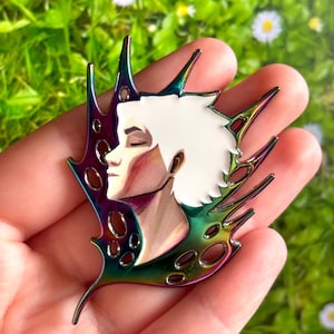 May include: A metal pin depicting a person's profile with white hair and closed eyes. The figure is set against an iridescent, multi-coloured background with a wing-like design. The pin has a smooth, polished finish.