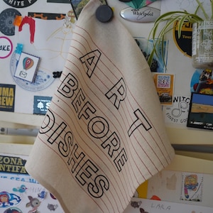 Puede incluir: Un paño de cocina beige con rayas rojas y las palabras "ART BEFORE DISHES" en letras negras grandes. El paño está colgado de un refrigerador cubierto de imanes y pegatinas.