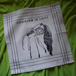Puede incluir: Un paño de cocina beige con una ilustración en negro de una mano sosteniendo un vegetal y el texto "COOKING IS LOVE". El paño tiene un diseño de borde a rayas y se muestra sobre una superficie verde.