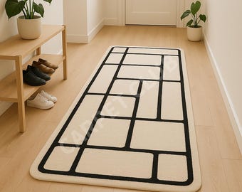 Puede incluir: Alfombra rectangular color crema con un diseño geométrico negro. La alfombra presenta una serie de rectángulos y cuadrados interconectados, creando un patrón abstracto moderno. La alfombra está colocada sobre un suelo de madera clara.
