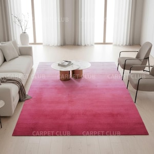 May include: A contemporary living room with a large, rectangular, ombre pink rug. The rug fades from a lighter pink to a deeper shade. The room includes a white sofa, two grey armchairs, and a round coffee table.