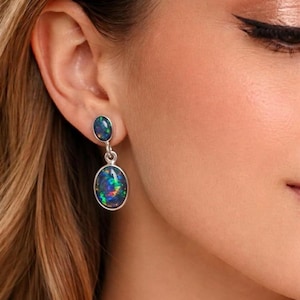 May include: Silver drop earrings featuring oval-shaped black opal stones with iridescent color flashes. The earrings have a small, round opal stone at the top. The jewelry is worn on a person's ear.