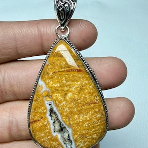 May include: A teardrop-shaped pendant with a silver-coloured bail. The pendant features a yellow and brown stone with a white and black fissure. The stone is framed by a silver-coloured, beaded edge.