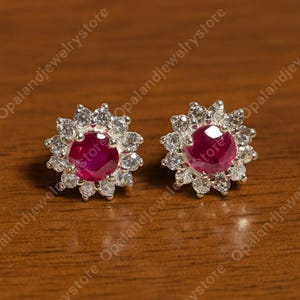 May include: A pair of stud earrings featuring a central, round, deep red gemstone surrounded by a halo of smaller, clear gemstones. The earrings are set in a silver-colored metal and are displayed on a wooden surface.