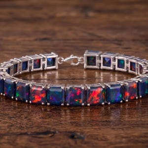 May include: A silver-toned link bracelet featuring a series of square-cut black opals. Each opal displays vibrant flashes of red, orange, and green color play within the dark stone. The stones are set in individual metal frames connected by hinges.
