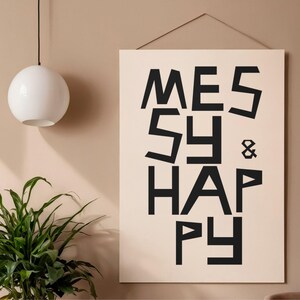 May include: A rectangular wall hanging featuring a light beige background with the black text "MESSY & HAPPY" printed in a bold, geometric, sans-serif typeface. The artwork is suspended by a thin string attached to the top edge of the frame.