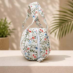 Pu&ograve; includere: Una pochette in tessuto arrotondata con base bianca caratterizzata da un motivo floreale di piccoli fiori e foglie rosse, blu e verdi. La borsa presenta un unico manico superiore e una chiusura a zip bianca verticale sul pannello frontale.