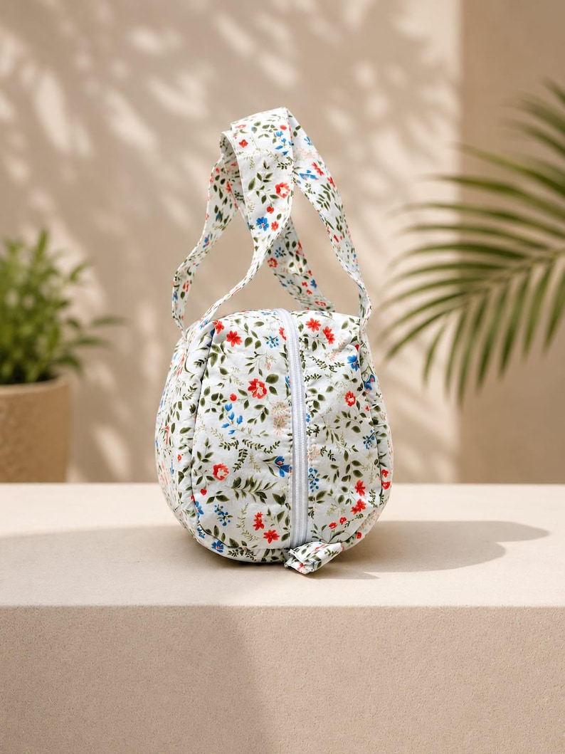 Pu&ograve; includere: Una pochette in tessuto arrotondata con base bianca caratterizzata da un motivo floreale di piccoli fiori e foglie rosse, blu e verdi. La borsa presenta un unico manico superiore e una chiusura a zip bianca verticale sul pannello frontale.