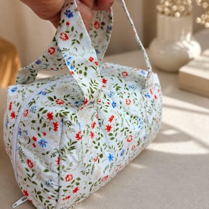 Pu&ograve; includere: Una piccola borsa cilindrica in tessuto con base bianca e un motivo ripetuto di minuscoli fiori rossi e blu e foglie verdi. La borsa include due manici in tessuto abbinati e una chiusura a zip bianca sul lato per l'apertura.