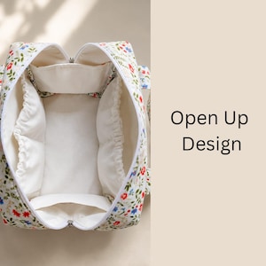 Pu&ograve; includere: Una borsa da viaggio trapuntata bianca con un colorato motivo floreale di fiori rossi, blu e verdi. La borsa &egrave; aperta e rivela un interno color crema con diverse tasche elasticizzate e una chiusura a zip. Il testo recita Open Up Design.