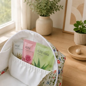 Pu&ograve; includere: Una borsa cosmetica in tessuto bianco con motivo floreale contenente tre maschere per il viso. Le maschere sono etichettate come Rice Brightening Mask, Rose Moisturizing Mask e Aloe Vera Soothing Mask, ognuna con illustrazioni botaniche.