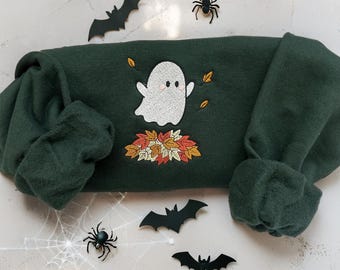 Può includere: Felpa verde scuro con un ricamo a forma di fantasma. Il fantasma bianco con guance rosa fluttua sopra un mucchio di foglie autunnali. Pipistrelli e ragni decorativi circondano la felpa, creando un tema di Halloween.