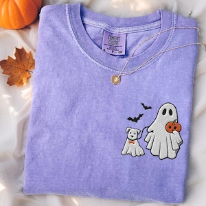 May include: A lavender Comfort Colors t-shirt with a Halloween-themed embroidered design. The design features a ghost holding a pumpkin, a dog with a bow tie, and two bats. A gold necklace with a pendant is also visible.