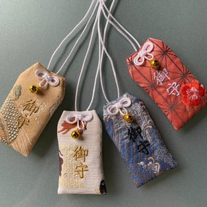 May include: Four rectangular fabric omamori amulets with white hanging cords and small gold bells. Each features embroidered patterns and the Japanese kanji characters for omamori. The colors include tan, white, blue, and coral with floral motifs.