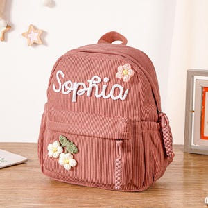 Może przedstawiać: Różowy plecak sztruksowy z imieniem "Sophia" wyhaftowanym białym pismem. Plecak posiada górny uchwyt, przednią kieszeń i aplikacje kwiatowe. W tle oprawiony obrazek i książki dla dzieci.
