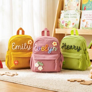 May include: Three corduroy backpacks in yellow, pink, and green, each personalized with a name in cursive lettering. The yellow backpack says "Emily", the pink says "Evelyn", and the green says "Henry". Each bag has a front pocket and a flower embellishment.