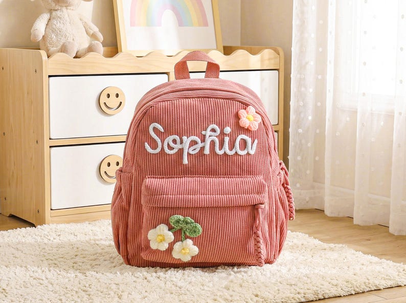 Op de afbeelding: Een oudroze rugzak van corduroy met de naam Sophia in witte schrijfletters geborduurd aan de bovenzijde. De tas heeft een voorvak met rits, versierd met twee gehaakte witte bloemen met groene blaadjes en een roze bloem aan de rechterbovenkant.