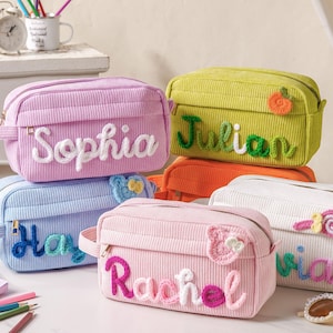 Può includere: Una collezione di trousse in velluto a coste personalizzate in vari colori, tra cui rosa, blu e verde. Ogni borsa presenta un nome ricamato in un colore a contrasto. Misure in centimetri.