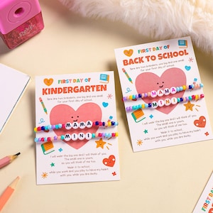 May include: Two bracelets with the words "MAMA" and "MINI" on cards that say "FIRST DAY OF KINDERGARTEN" and "BACK TO SCHOOL". The cards have a pink heart and school-themed illustrations. A pink pencil sharpener and pencils are in the background.