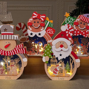 以下が含まれることがあります： 木製のクリスマス飾りで、雪だるま、ジンジャーブレッドマン、サンタクロースが特徴で、それぞれお祝いのデザインと内部照明が施されています。雪だるまのベースには「Olivia」という名前が書かれています。サンタクロースのベースには「Louis」という名前が書かれています。