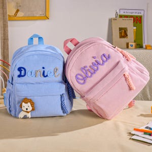 Op de afbeelding: Twee rugzakken van corduroy, een lichtblauw en een lichtroze. De blauwe rugzak heeft de naam Daniel in blauw schrift en een kleine leeuwenpatch. De roze rugzak heeft de naam Olivia in paars schrift. Beide hebben handvatten aan de bovenkant.
