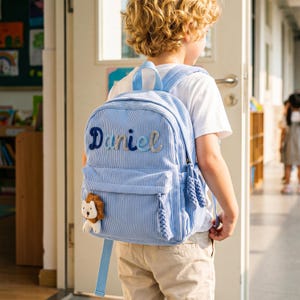 Op de afbeelding: Een lichtblauwe rugzak van corduroy met een verticale streeptextuur en een voorvak. De naam Daniel is in donkerblauw en lichtblauw cursief geborduurd op de voorzijde. Een kleine pluche leeuwbedel is bevestigd aan de ritssluiting.