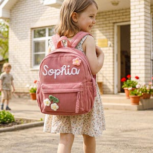 Op de afbeelding: Een oudroze rugzak van corduroy met de naam Sophia in witte schrijfletters op het voorvak. De tas is versierd met een kleine gehaakte bloemapplicatie aan de bovenkant en twee witte gehaakte bloemen met groene blaadjes aan de onderkant.