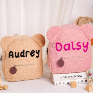 May include: Two teddy bear-shaped backpacks, one tan and one pink, with the names "Audrey" and "Daisy" embroidered on them. Each backpack has a zippered front pocket and a small, round charm. The backpacks are sitting on a white surface.