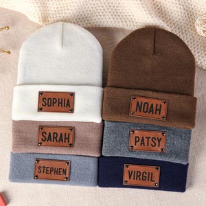 Puede incluir: Seis gorros personalizados en varios colores: blanco, marrón, gris y azul marino. Cada gorro tiene un parche de cuero marrón con un nombre: Sophia, Sarah, Stephen, Noah, Patsy y Virgil. Los gorros están apilados.