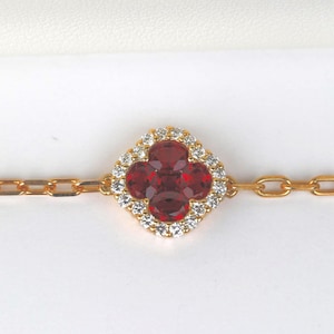 May include: Gold-toned bracelet featuring a four-leaf clover pendant with four red gemstones at the center, surrounded by a halo of small, clear stones. The bracelet is set against a white background.