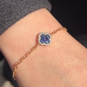 Puede incluir: Pulsera dorada con un colgante cuadrado con piedras azules y pequeñas piedras blancas. La pulsera tiene un diseño de cadena fina y se lleva en la muñeca.