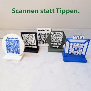 Könnte beinhalten: Eine Sammlung von QR-Code-Display-Ständern in verschiedenen Formen und Farben. Die Ständer umfassen einen weißen runden Ständer mit blauem QR-Code, einen schwarzen rechteckigen Ständer, einen weißen rechteckigen Ständer, einen grünen sechseckigen Ständer und einen blauen geometrischen Ständer.